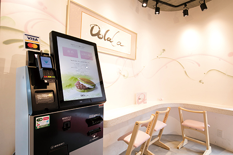 有機茶生どら専門店「Dolala」（東京）。茶室を思わせるこだわりの空間にも溶け込むモダンなフォルム。高解像度の画面が商品の魅力を一層引き立てる。スタンダードタイプにキャッシュレス機能を追加 スタンダードタイプ（w495×d320×h1510㎜、重量:105kg）、キャッシュレスタイプ［スタンド型］（w495×d320×h1510㎜、重量:60Kg）、キャッシュレスタイプ［カウンタートップ型］（w495×d340×h675㎜、重量:30Kg）