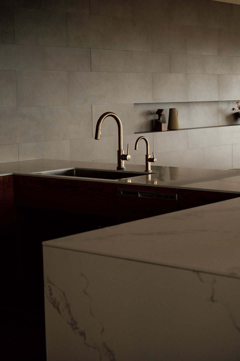 メイン写真の右隣にあるキッチン。クォーツ製のコの字型カウンターは柄が連続するように絶妙にそろえている。オーク無垢材の床、カウンターのクォーツ、ゴールドの水栓、壁面のタイルの調和が美しい。壁面は構造を隠すためにふかし、奥行き180㎜の棚を設えた